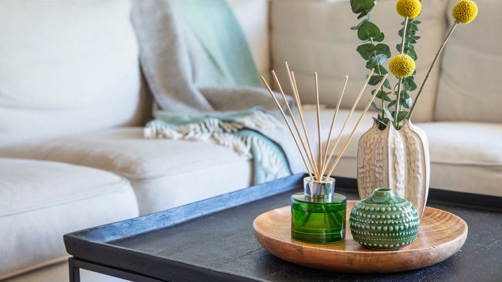 A beige sofa with a green and gray throw blanket, and a black coffee table with a wooden tray, a green diffuser, ceramic vases and greenery.